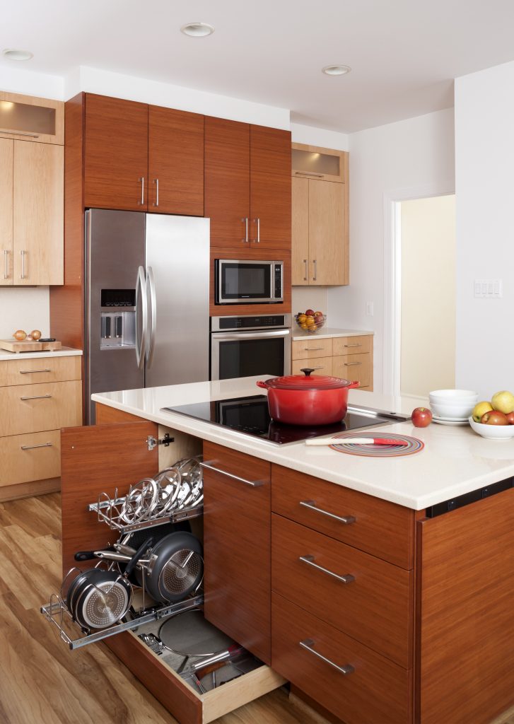 A clean, well-designed kitchen with a mix of light brown and red-brown cabinets. A red-brown cabinet built into the kitchen island is open, revealing a pull-out drawer with neatly arranged pans and lids. An orange enamel pot sits on an electric stovetop on the island.