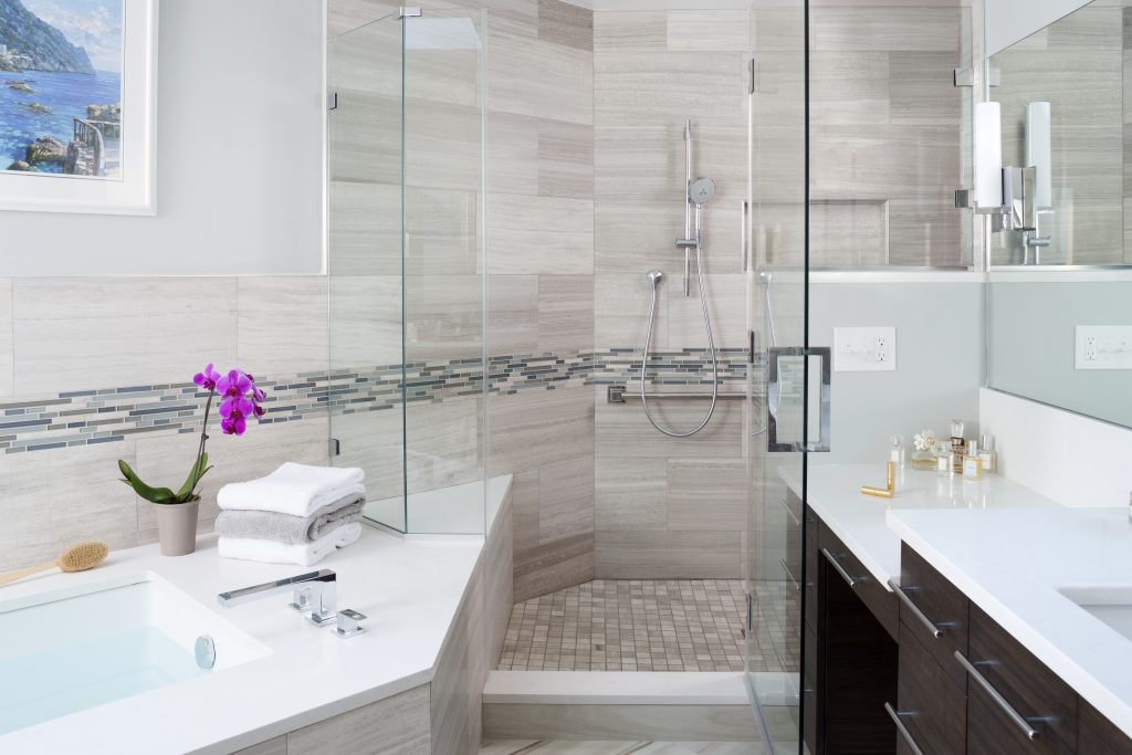 A large shower with a glass door, adjustable hand-held shower head, and a stability bar stands in the corner. On either wall of the corner are white countertops with sinks. On the counters are skin-care bottles, white and grey towels, and a purple flower.