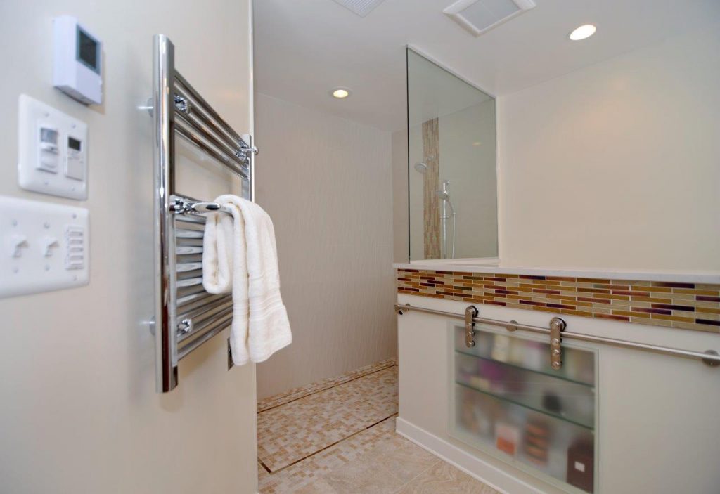 A white towel hangs from a towel warmer, next to light switches and a control panel. Beyond the towel is a shower with a glass door, brown and white tiles, and handheld shower head. A counter on the side of the room contains a pebbled glass medicine cabinet and metal railing.